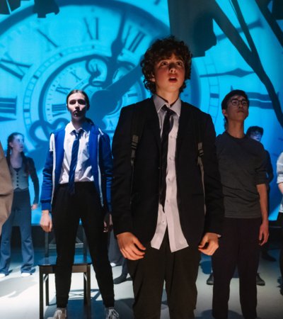 Middle School pupils on stage performing in A Monster Calls with a projection of a clock on the wall