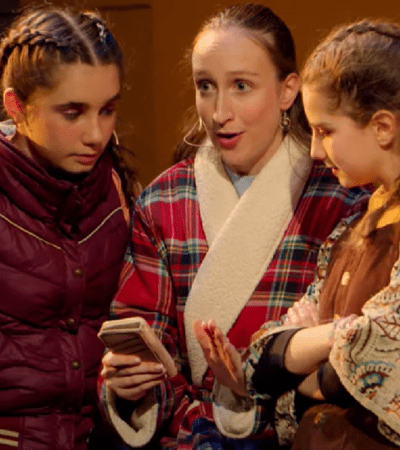 Alice in a dressing gown holding a phone on stage in a play with her theatre group