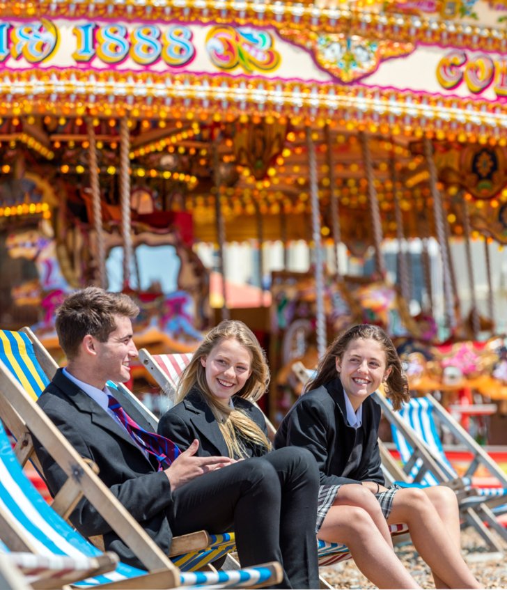  Pupils in their uniform sitting on Brighton Beach in deck chairs in front of a carousel 