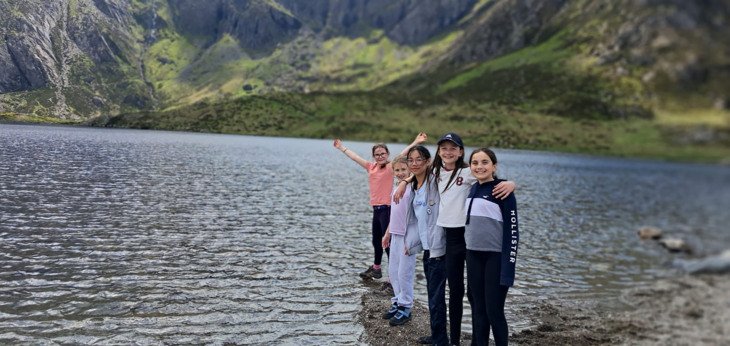  Prep-School pupils enjoying their school trip standing on the shore of a lake with mountains in the background 