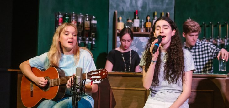  Pupils in the production of Two sitting on red stools performing on the guitar and singing into a microphone in front of the bar 