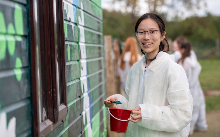  Pupil on overalls with a bucket of paint and a brush painting a mural for Make A Difference Day 