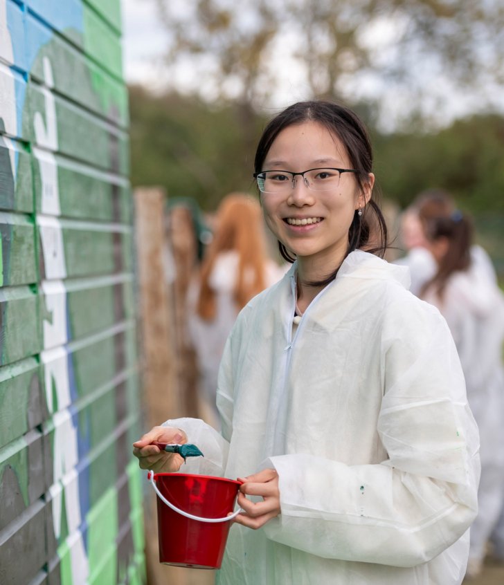  Pupil in painting overalls holding a bucket of paint and painting a mural on a wall in a park in Brighton 