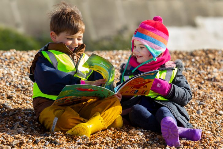  Nursery pupils sitting on the stones on the beach enjoying reading a book 