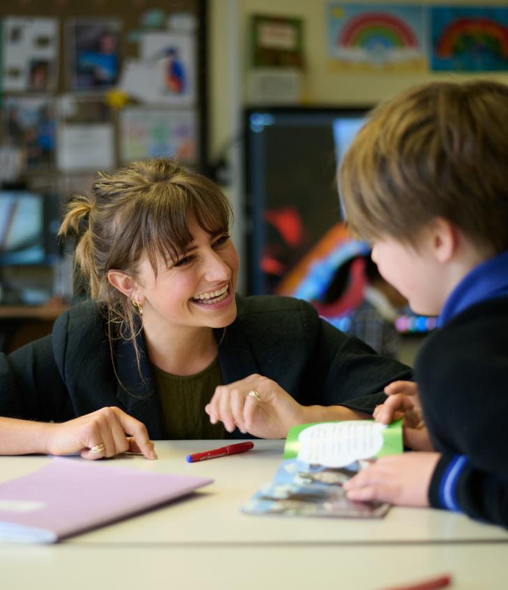  Pre-Prep pupil sitting at a desk and reading a book with their teacher in the classroom 